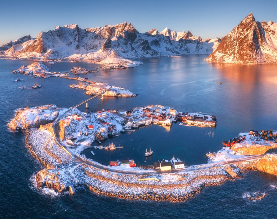 Aerial view of Reine and Hamnoy at sunset in winter. Amazing Lofoten islands, Norway. Panoramic landscape with blue sea, snowy mountains, rocks, village, buildings, rorbuer, road, bridge, sky.Top view